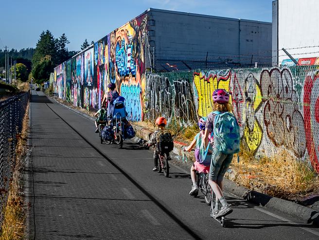 a family riding bikes and scooter by a graffitied wall