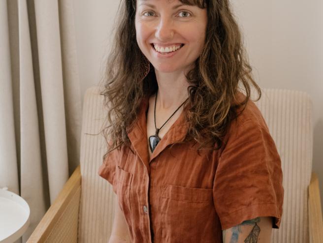 Portrait of Cedra Woods seated in a light-coloured armchair, smiling at the camera. She has wavy brown hair with bangs and a small nose ring, and is wearing a rust-coloured short-sleeve linen outfit. Her forearms show visible tattoos, and the setting is a calm indoor space with soft neutral tones and curtains in the background.